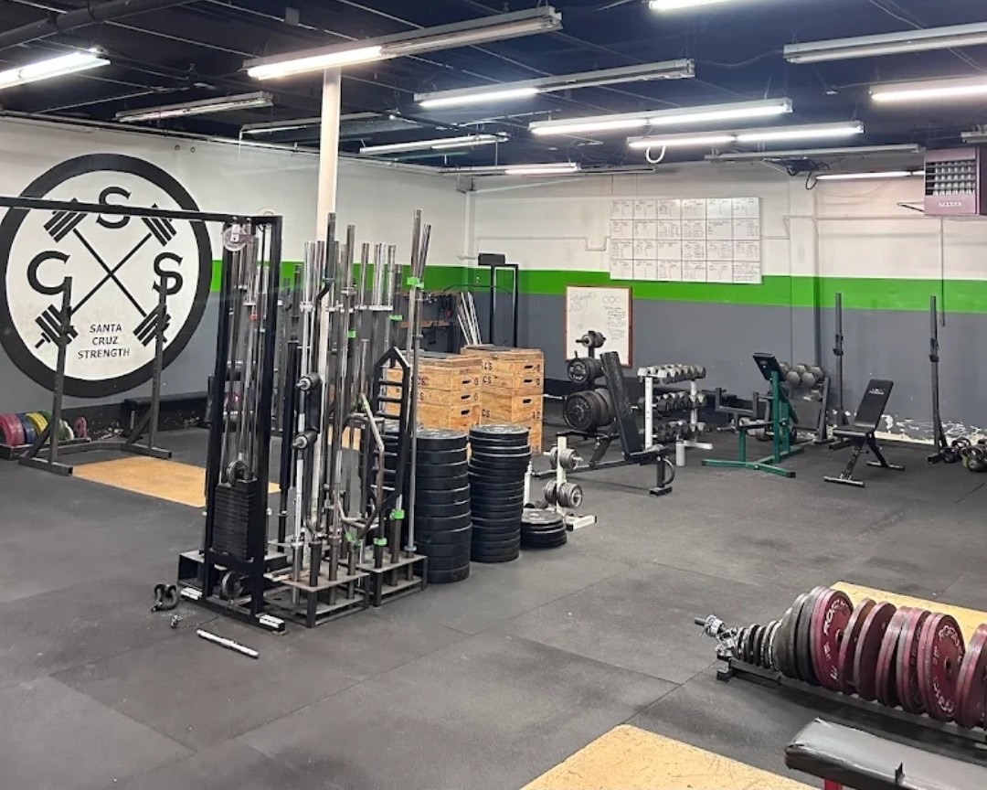 The Santa Cruz Strength training floor: the painted SCS wall seal, bumper and iron plates stored at floor level, power racks along the wall, and wooden lifting platforms on the open floor